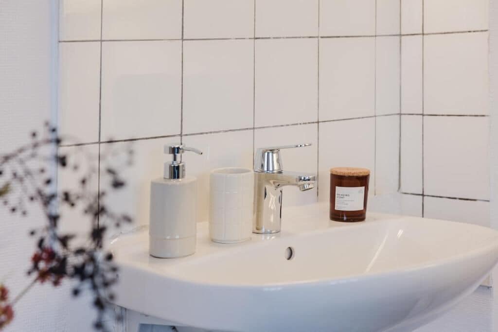 Une salle de bain avec un lavabo blanc, deux robinets, un gobelet blanc et un pot de savon.