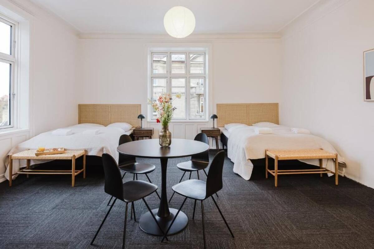 Habitación de hotel con dos camas, una mesa de comedor con sillas, un florero con flores y una ventana con vistas.