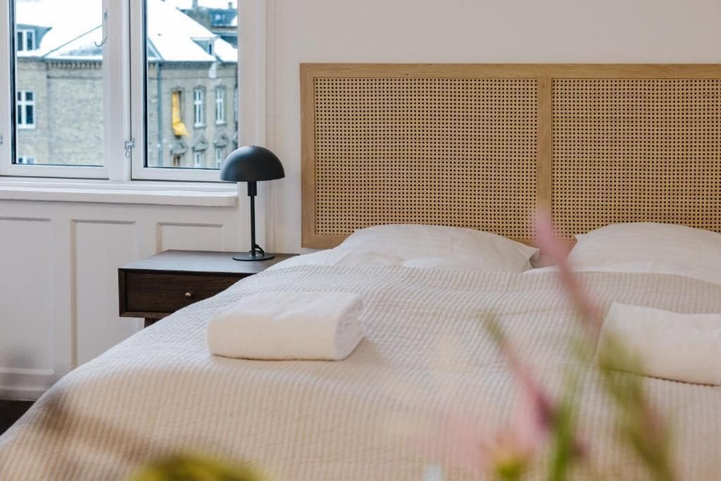 Ein ordentlich bezogenes Bett mit Kopfteil, einer Nachttischlampe und einem Fenster mit Blick auf Gebäude.