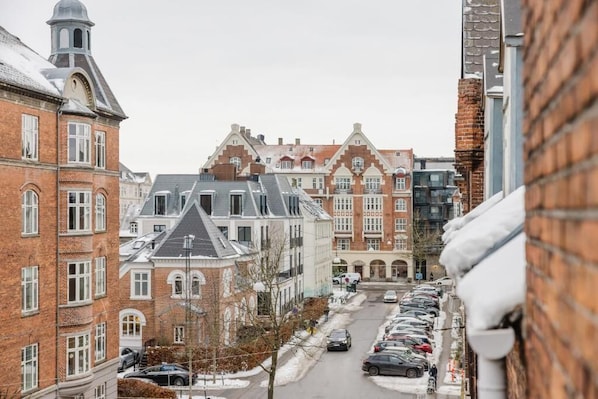 Vista de la calle con una fila de coches aparcados, un edificio de ladrillo con una torre y una acera cubierta de nieve.