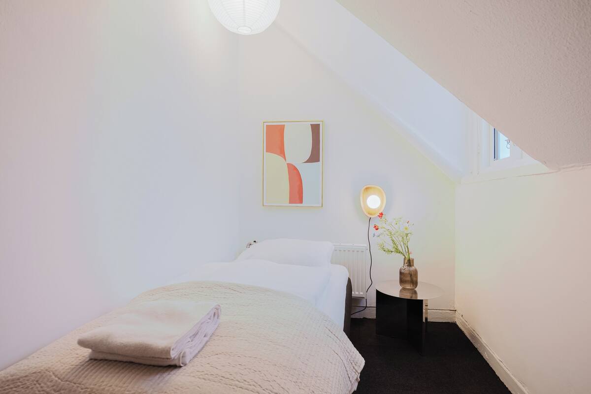 Un dormitorio con una cama, una mesita de noche con una lámpara, un florero con flores y una obra de arte en la pared.