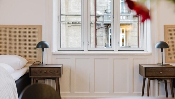 Hotellihuone, jossa on sänky, kaksi yöpöytää, ikkuna, josta on näkymä rakennuksiin, ja lamppu kummallakin pöydällä.