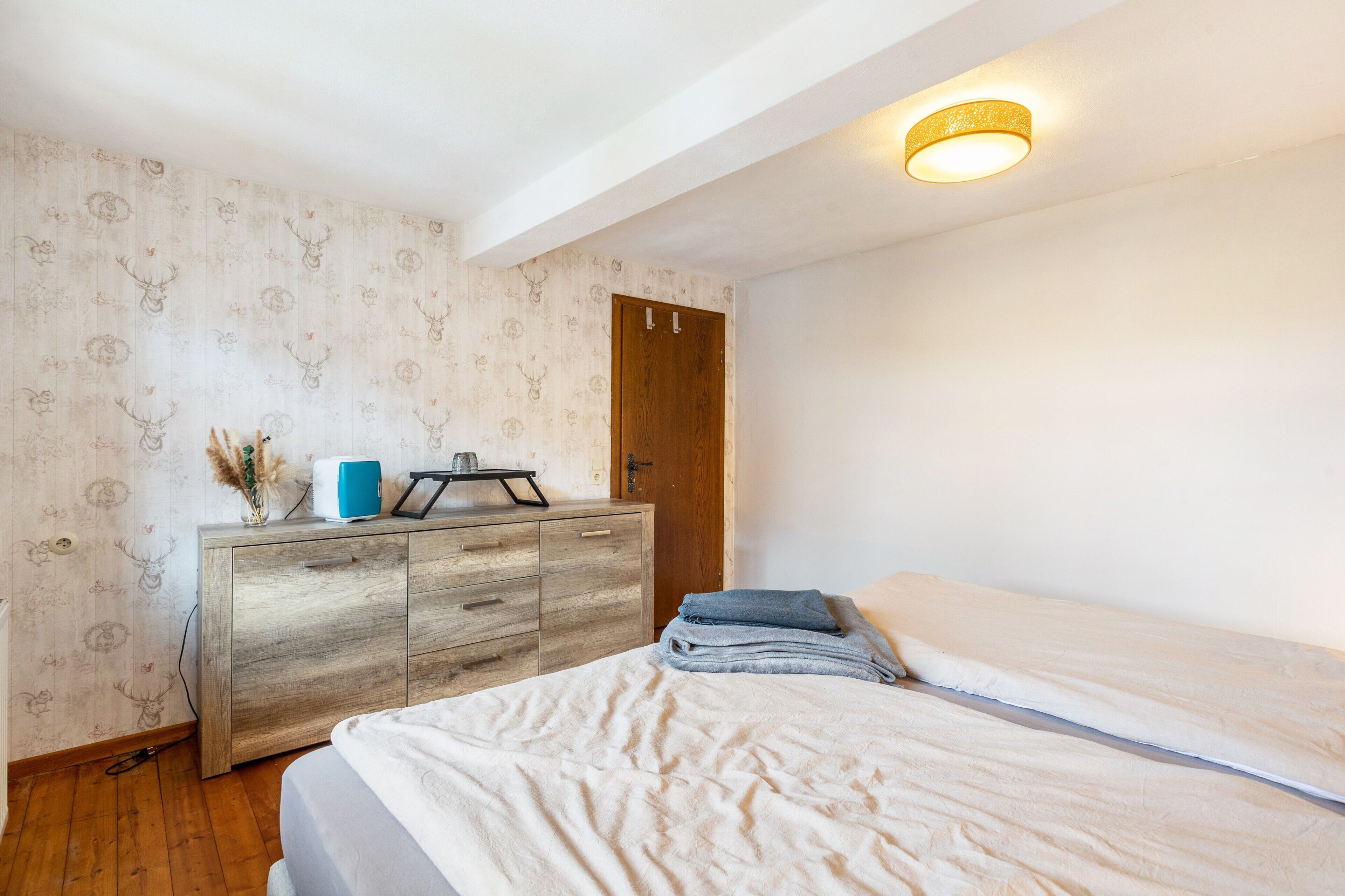 A bedroom with a wooden dresser, a bed with a white comforter, a wooden door, and a ceiling light fixture.