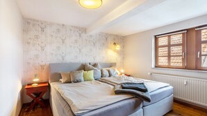 Un dormitorio con una cama, lámparas de noche, una ventana con contraventanas de madera y papel tapiz estampado.