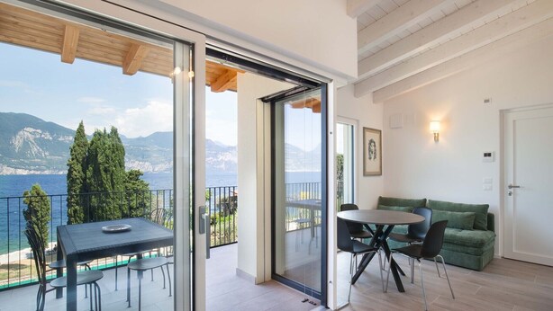 A balcony with a table and chairs, a green sofa, and a door leading to a room with a view of the sea and mountains. - Pèler Apartment: Modern Elegance, Stunning Views and Comfort in Brenzone (Castelletto)