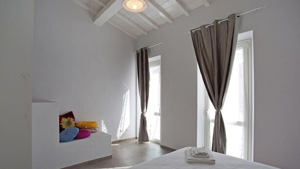 Quarto com cama, cortinas, janela e prateleira na parede com almofadas.