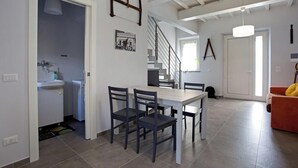 Uma sala de jantar com mesa e cadeiras, uma cozinha ao fundo, uma escada e uma porta com um casaco pendurado.