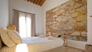 A bedroom with a stone wall, a bed with white linens, a bedside lamp, and a window with curtains.