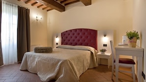 A bedroom with a red upholstered headboard, a bed with a beige bedspread, a bedside table with a lamp, a chair, and a small potted plant. - Tenuta San Giovanni Grapefruit (Lucca)