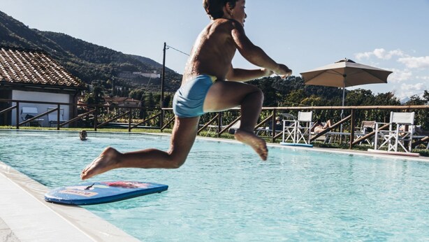A person jumping into a swimming pool with a blue kickboard, surrounded by lounge chairs and umbrellas. - Tenuta San Giovanni - Camomilla Apartment (Lucca)