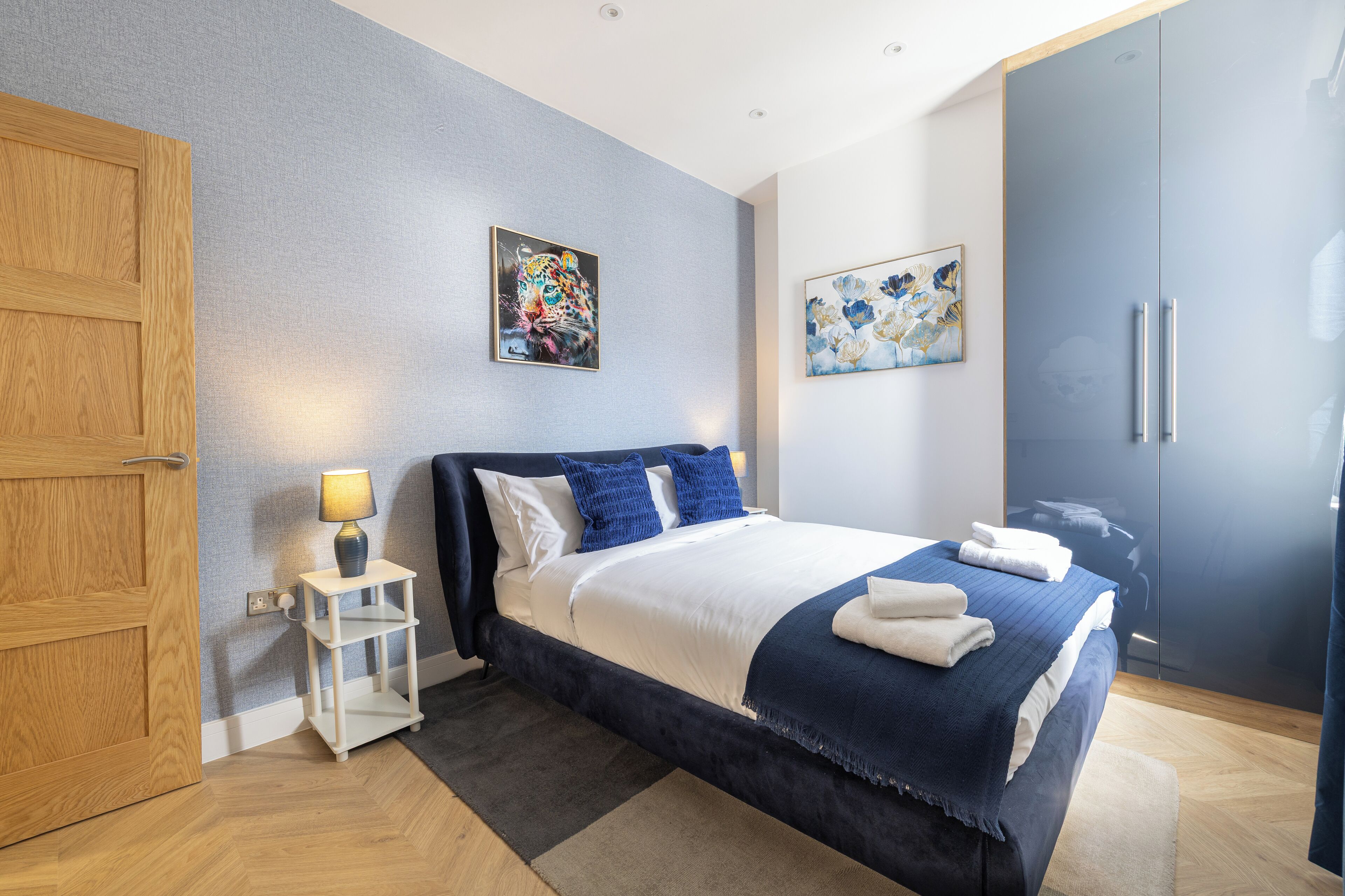 Quarto moderno com uma cama grande, uma porta de madeira, um criado-mudo com abajur e duas pinturas penduradas na parede.