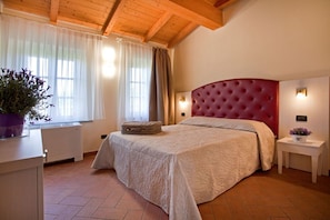 A bedroom with a large bed, wooden ceiling beams, a window with curtains, a bedside table with a lamp, and a potted plant. - Tenuta San Giovanni - Salvia Apartment (Lucca)