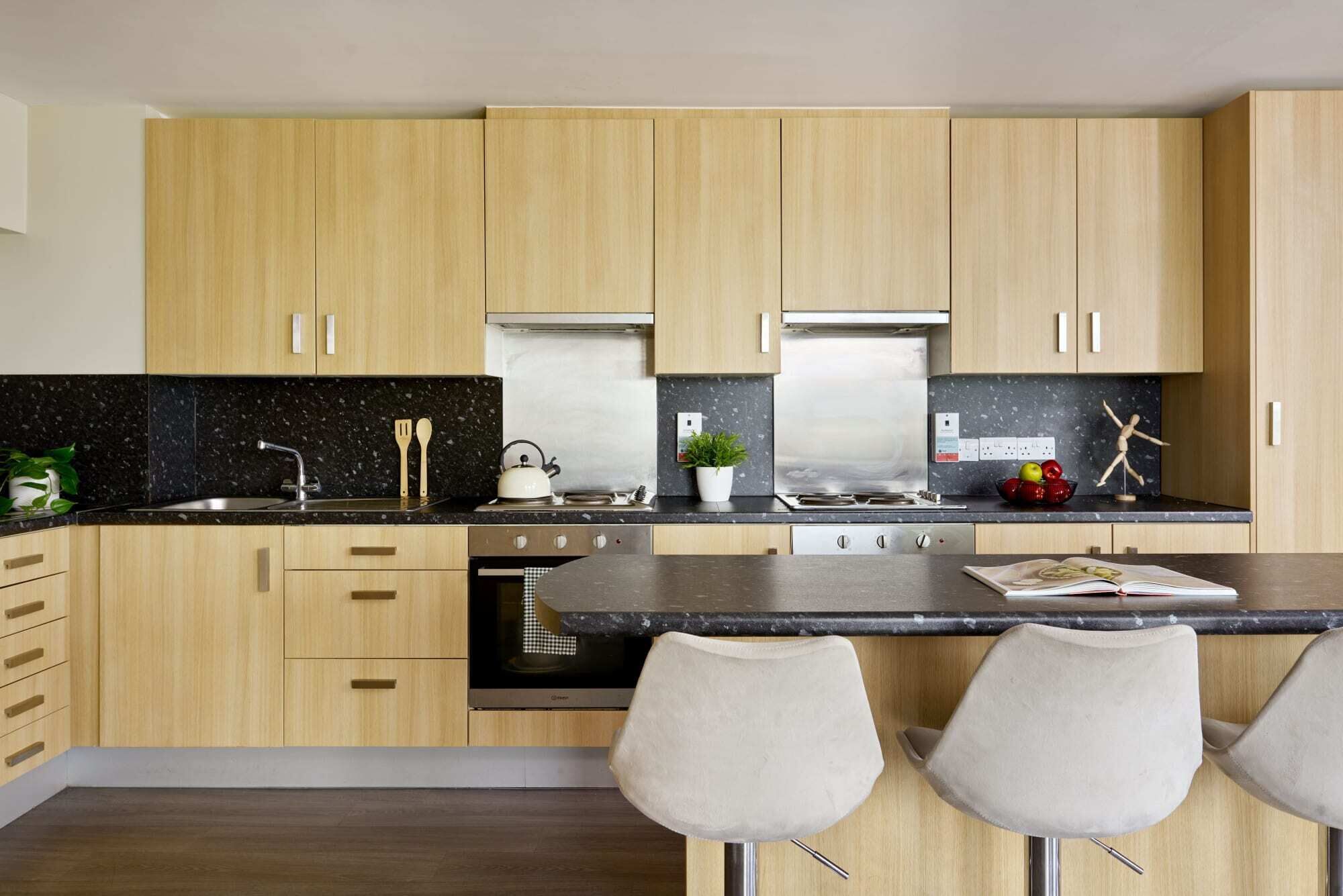 A modern kitchen with wooden cabinets, a black countertop, stainless steel appliances, and a bar area with two chairs.