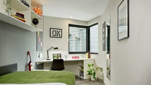 A modern bedroom with a bed, desk, and chair, a window with blinds, and a wall-mounted shelf with books and decorative items. - For Students Only Ensuites in Coventry (Coventry)