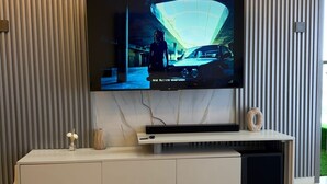 A modern living room with a large flat-screen TV mounted on a wall with a marble design. There is a white entertainment unit below the TV, and a round marble table in the foreground. - Urban Nest Westlands – Infinity Pool, Gym & Scenic Views close to major malls (Nairobi)