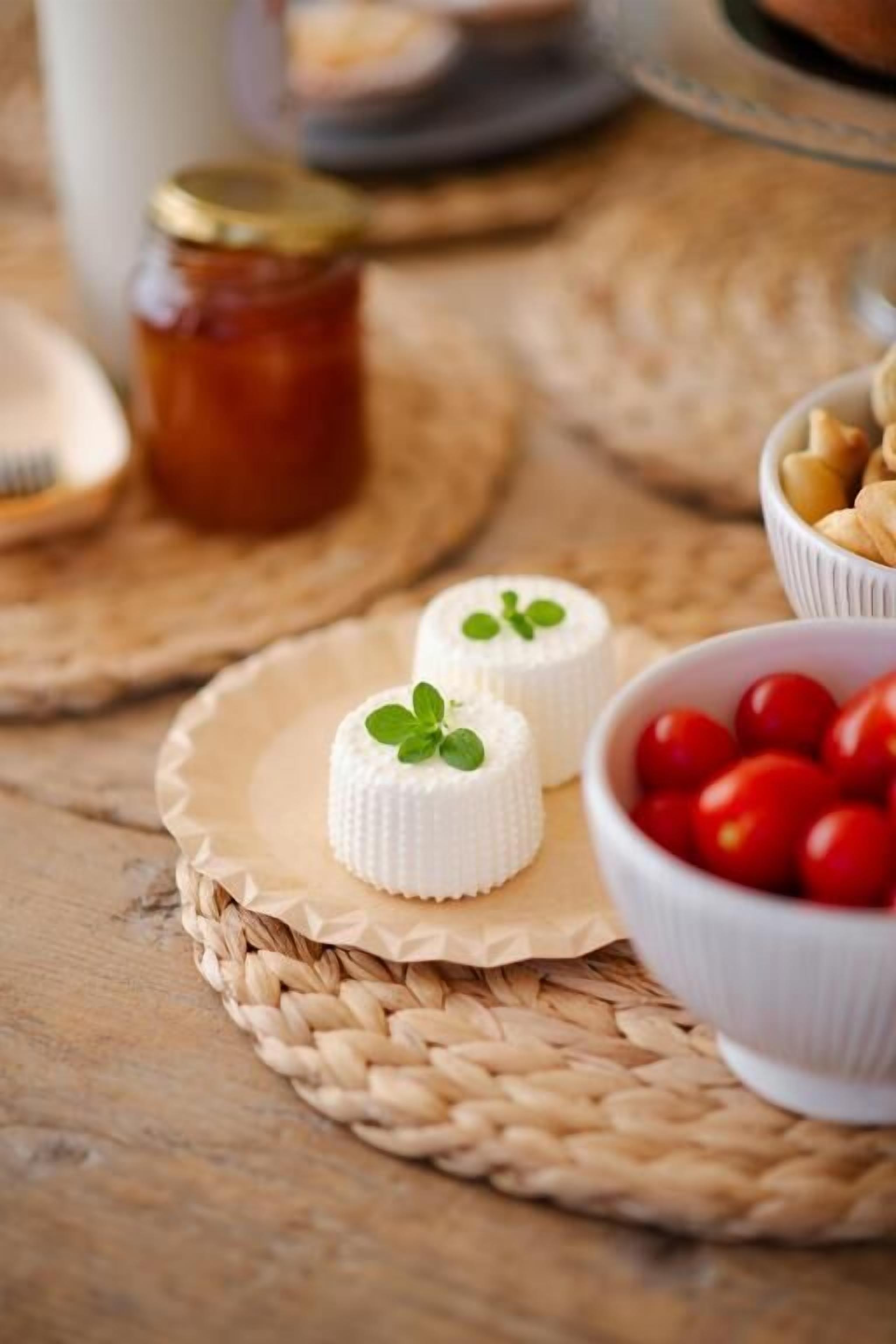 Dos pequeños quesos blancos con hojas verdes sobre una estera tejida, un cuenco de tomates cherry y un tarro de miel al fondo.