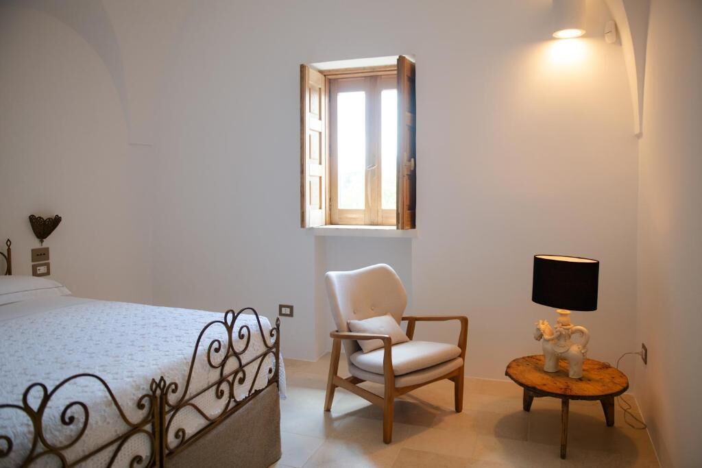 A bedroom with a bed, a chair, a small table with a lamp, and a window with curtains.