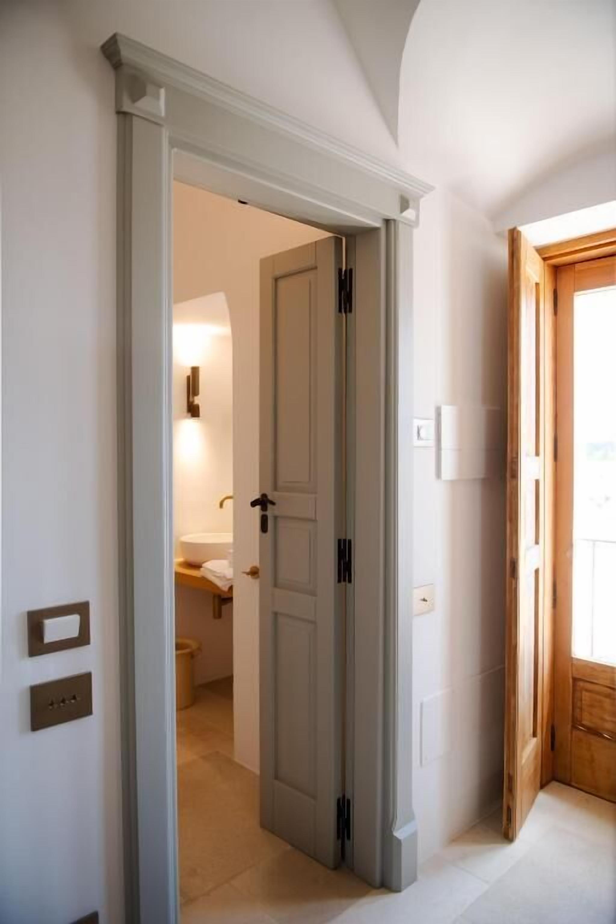 Un couloir avec une porte donnant sur une salle de bain. Le couloir a un sol et des murs de couleur claire, et il y a un cadre de porte en bois sur le côté droit.