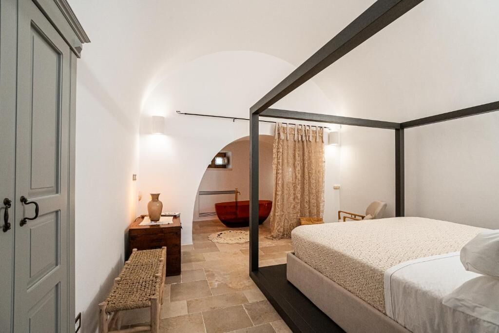 A bedroom with a four-poster bed, a small bedside table, a bench, and a red bowl in the background.