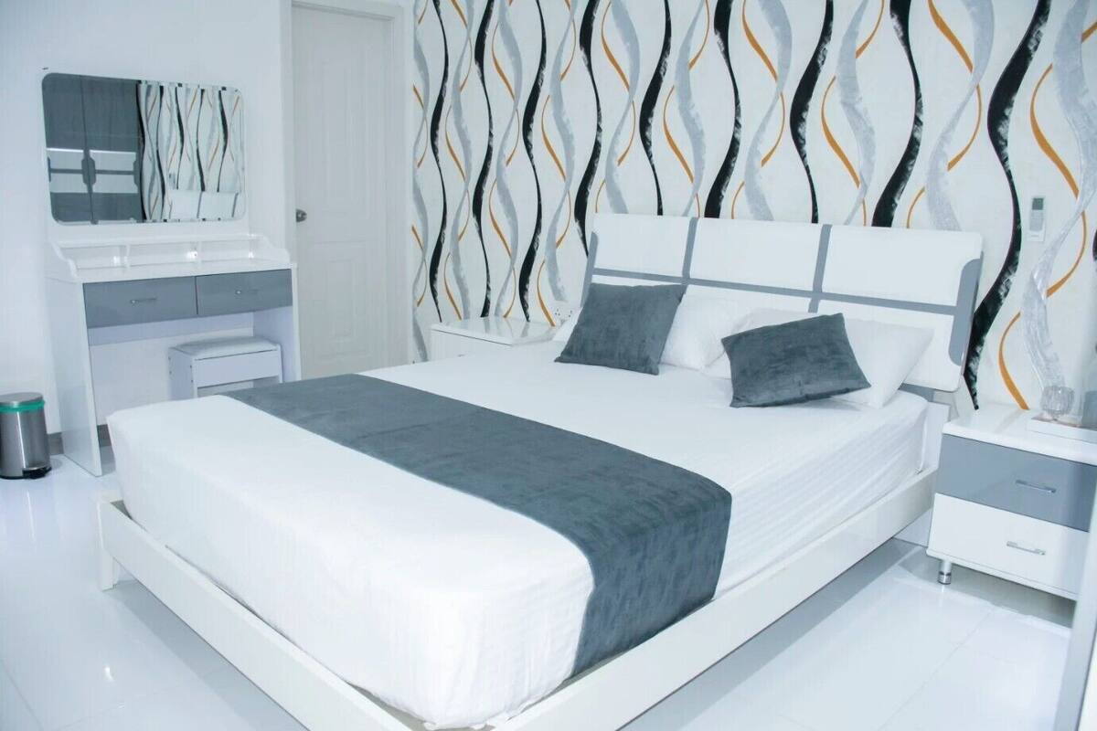 Una camera da letto moderna con un letto bianco, cuscini grigi, una coperta grigia, un comodino bianco e un tavolino di fianco al letto.