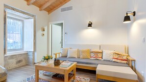 A cozy living room with a wooden ceiling, a sofa with patterned cushions, a coffee table, and a window with curtains. - Vinha Da Touça T1 Holiday Home with Mountain View, Wi-Fi and Air Conditioning (Soajo)