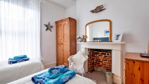 Hotellihuone, jossa on kaksi sänkyä, puinen vaatekaappi, valkoinen nojatuoli, takka ja sen yläpuolella peili.