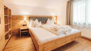 Una cama de madera con ropa de cama blanca, una mesita de noche, una lámpara de pared y una ventana con cortinas.