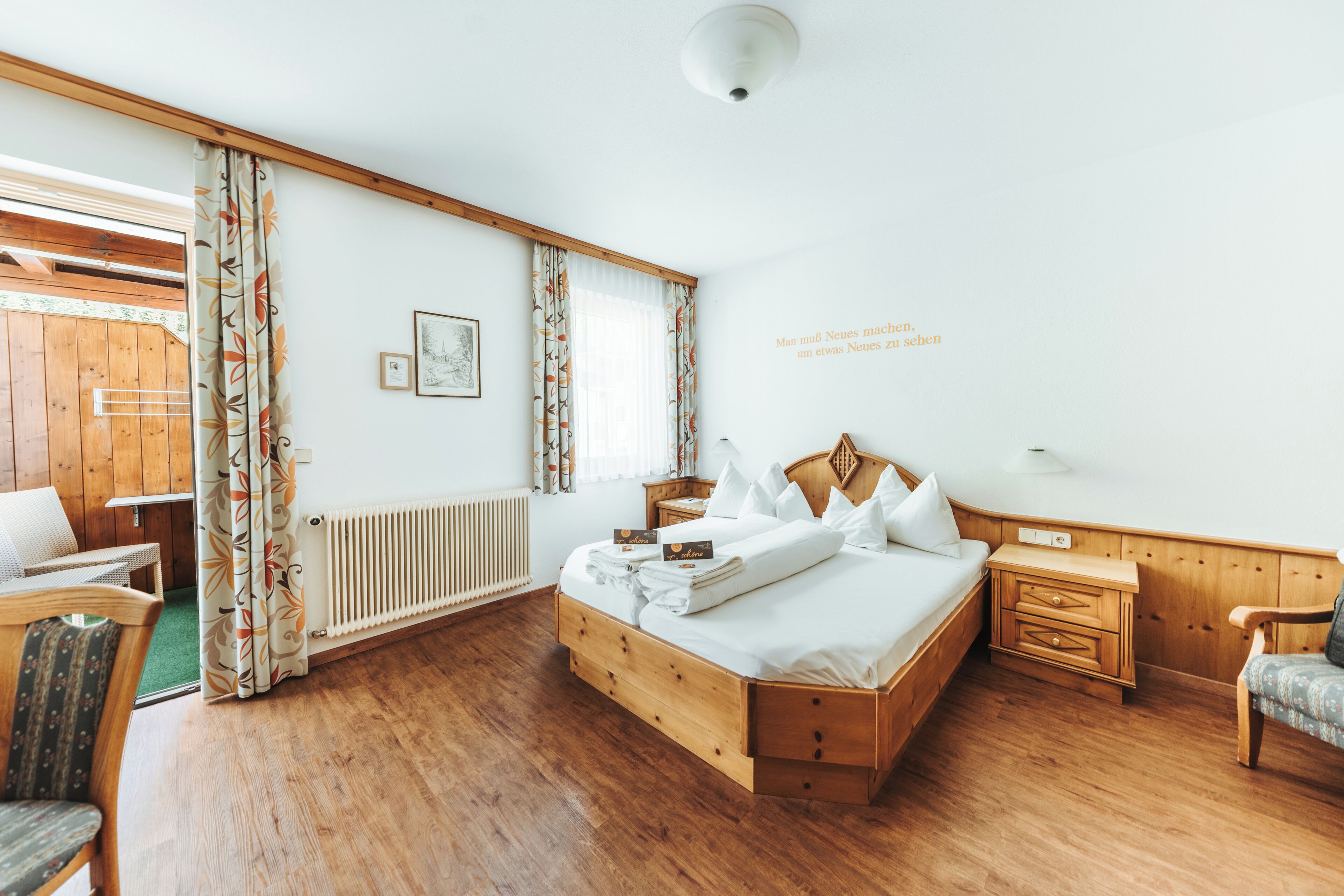 Una stanza con un letto di legno, un comodino in legno, una sedia, un tavolo e una finestra con tende.