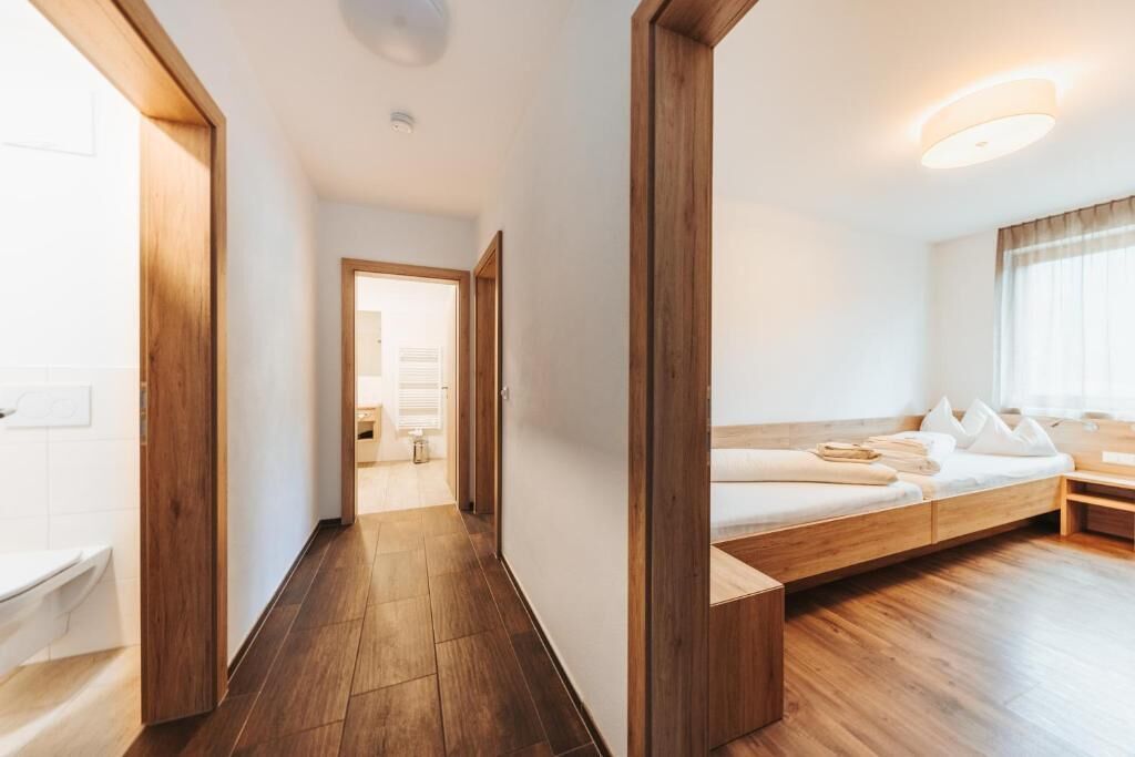 A modern hotel room with a wooden floor, a built-in bench with pillows, a bedside table, and a view of the hallway.