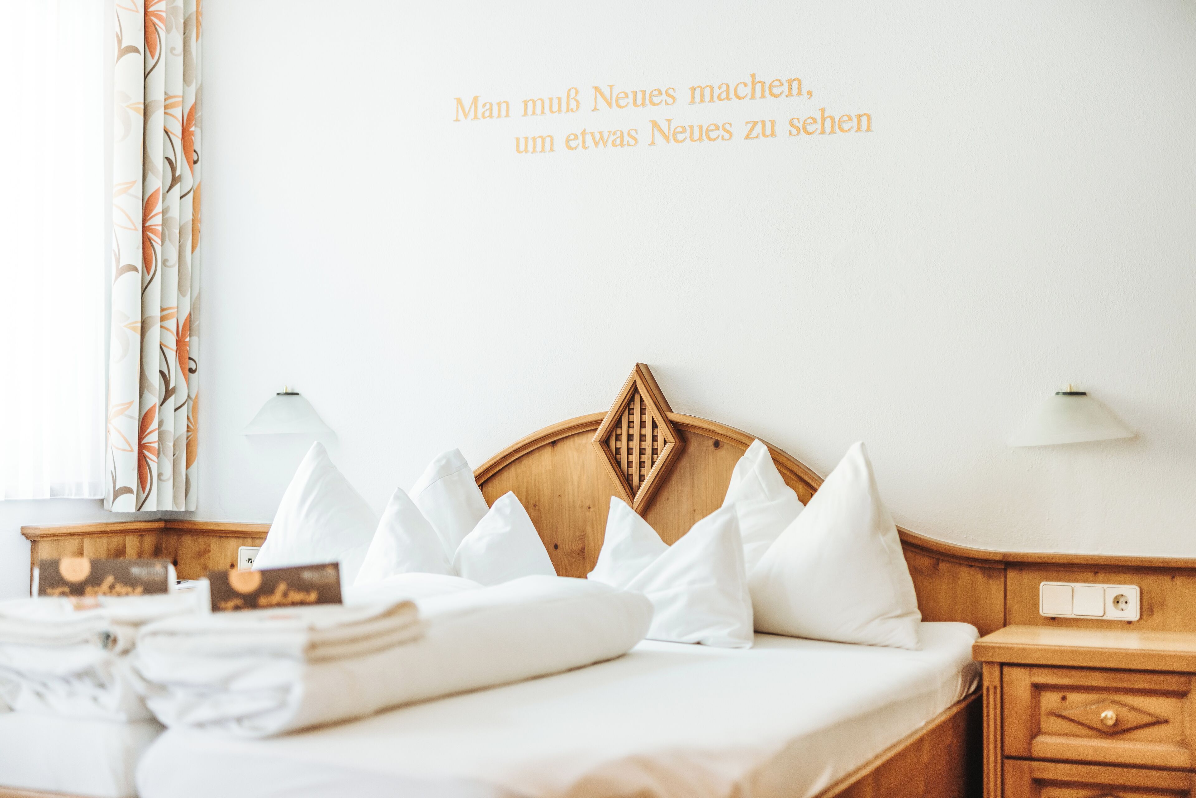 Ein ordentlich bezogenes Bett mit weißen Leinen und Kissen, ein hölzernes Kopfteil und eine Wand mit einem Text auf Deutsch.