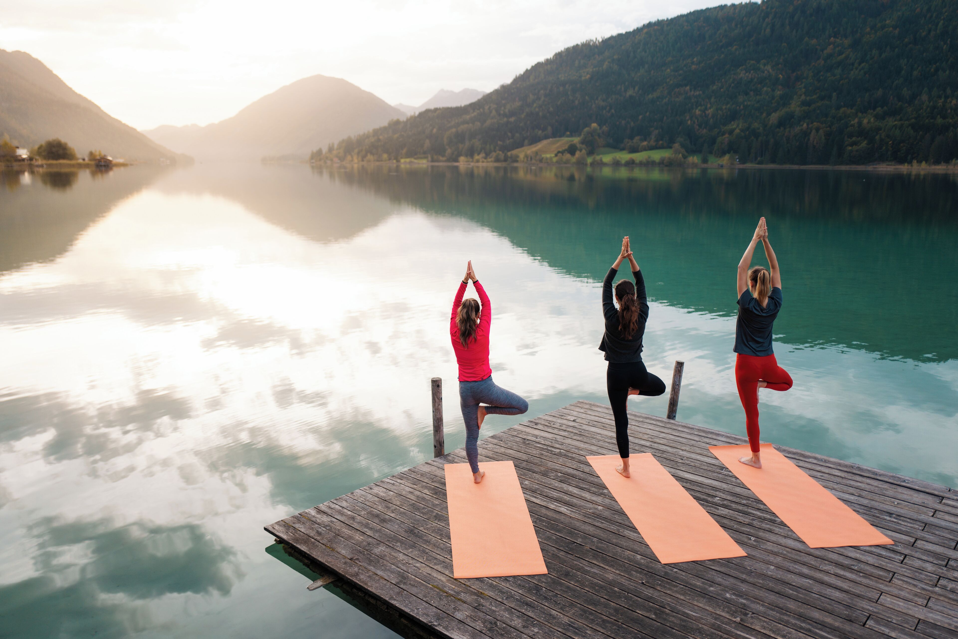 Tres personas practicando yoga sobre un muelle de madera frente a un lago tranquilo con montañas al fondo.