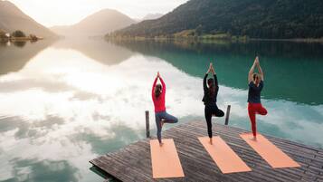 Três pessoas praticando yoga em um píer de madeira sobre um lago calmo, com montanhas ao fundo.
