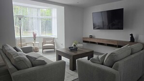 A modern living room with a sofa set, a coffee table, a TV mounted on the wall, and a window with blinds. - Woodside House (Forres)