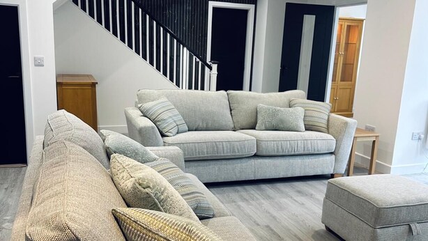 A modern living room with a grey sectional sofa, a matching ottoman, and a staircase with black railings. - Woodside House (Forres)