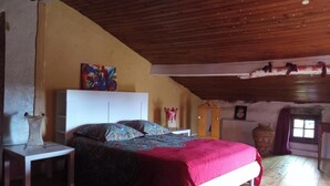 A bedroom with a sloped ceiling, a bed with a red cover, a white nightstand, a colorful abstract painting, and a wooden cabinet.