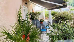 Un patio techado con mesa y sillas, rodeado de plantas en macetas y vistas al exterior.