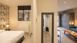 Un dormitorio moderno con cama, radiador, televisor montado en la pared y puerta que da a una sala de estar con sofá y lámpara.
