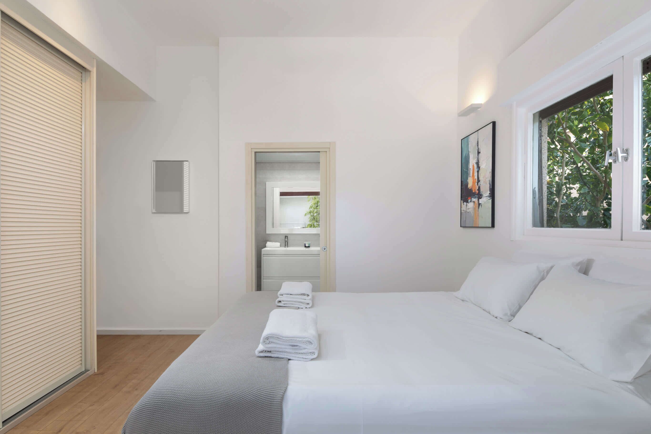 Una habitación de hotel moderna con una cama grande, un ventanal con vistas a zonas verdes, un baño con espejo y lavamanos, y una puerta que da a otra habitación.