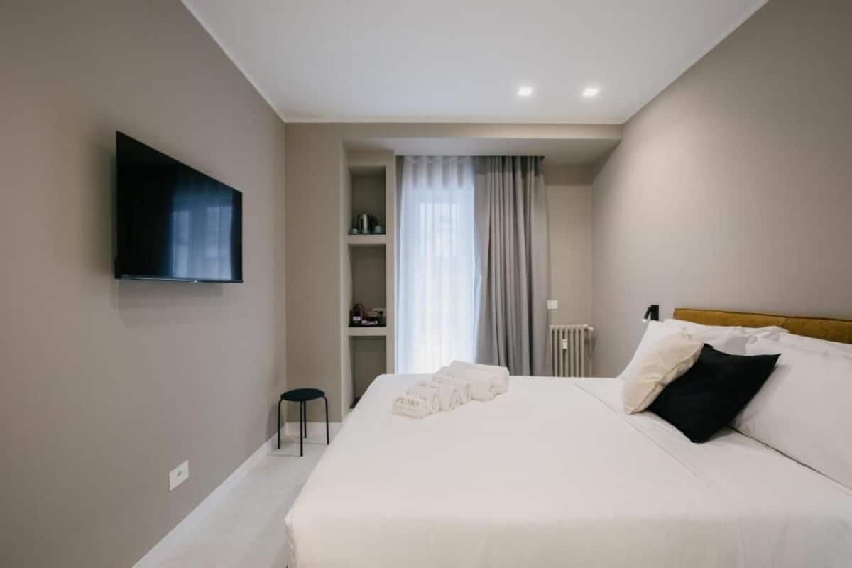 Une chambre d’hôtel moderne avec un grand lit, un téléviseur à écran plat fixé au mur, un petit tabouret et une fenêtre avec des rideaux.