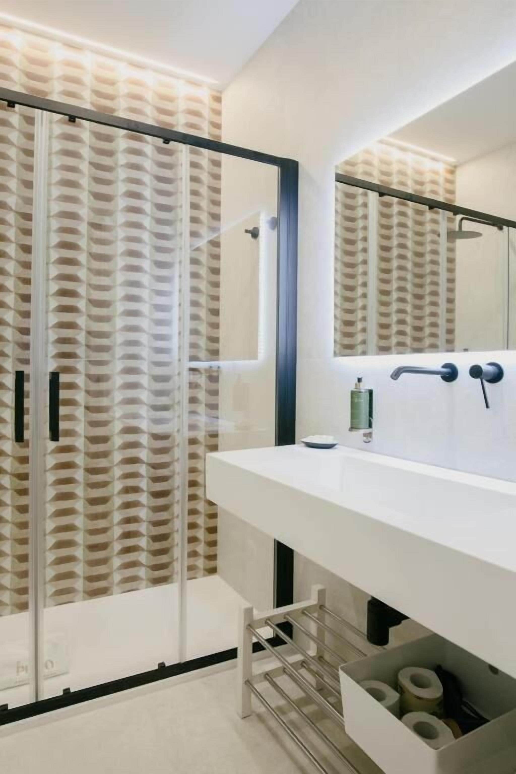 Un baño moderno con tocador blanco, un espejo grande y una zona de ducha con puerta de vidrio decorativa.