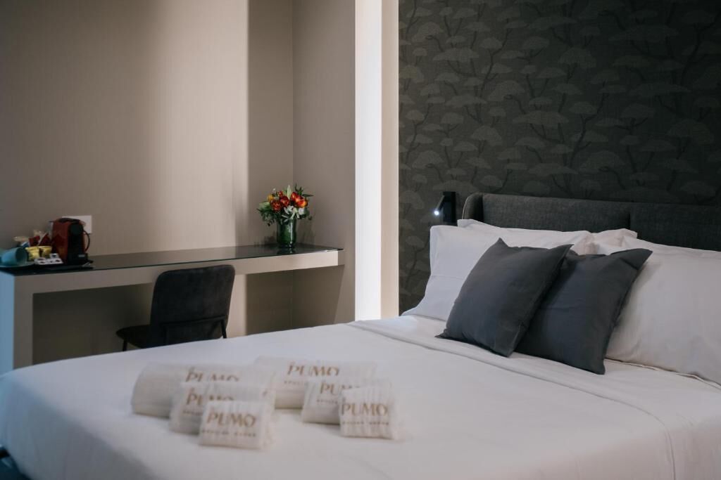 A hotel room with a bed, pillows, a desk, a chair, a vase with flowers, and a wall with a leaf pattern.