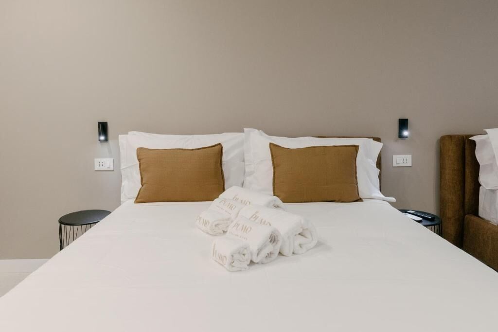 Cama bem arrumada com travesseiros brancos e castanhos, uma toalha dobrada e duas mesas de cabeceira com abajures pretos.