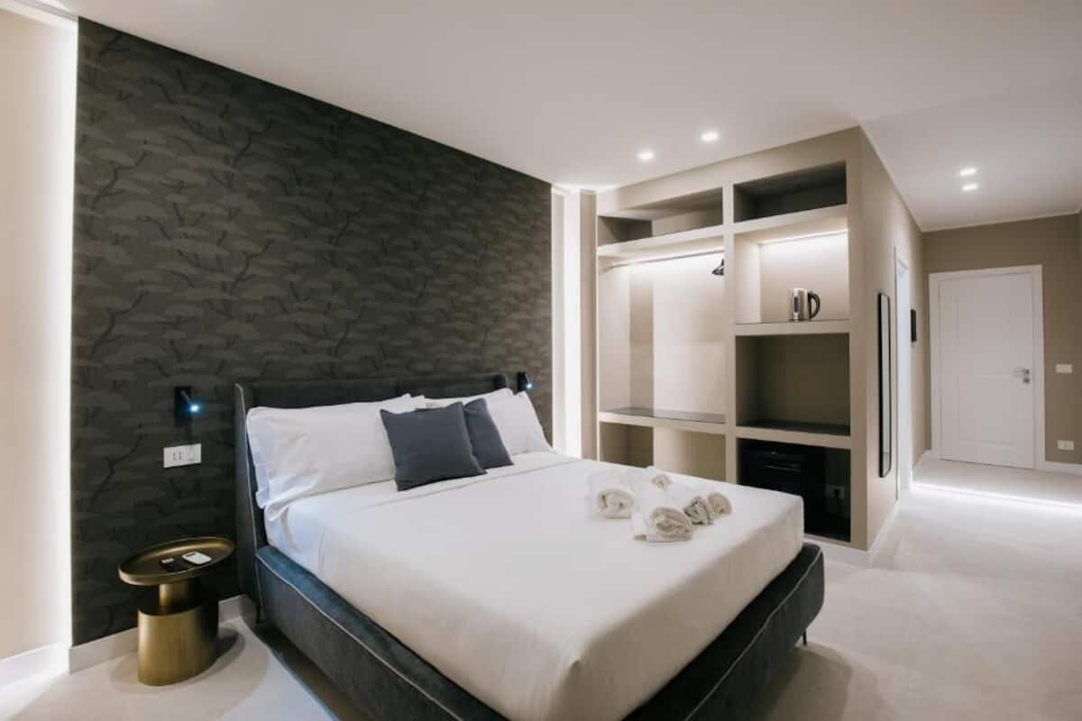 Une chambre moderne avec un mur texturé de couleur sombre, un lit bien fait avec du linge de lit blanc, une table de chevet avec une lampe et des étagères intégrées avec un miroir et des rangements.