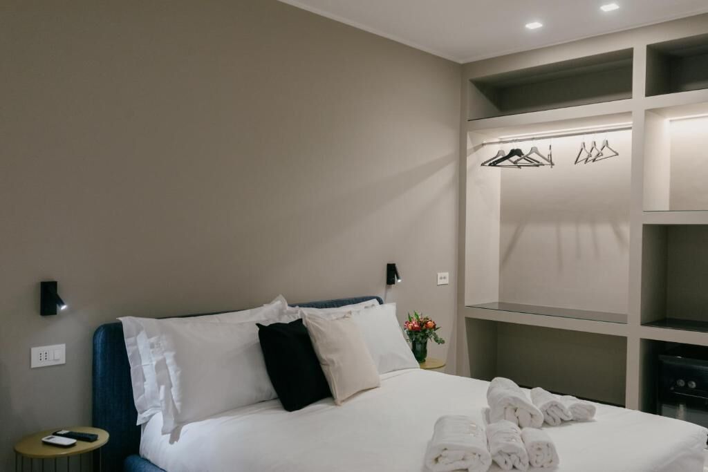 A hotel room with a bed, a bedside table with a phone, a wall-mounted light, a closet with hangers, and a vase with flowers.