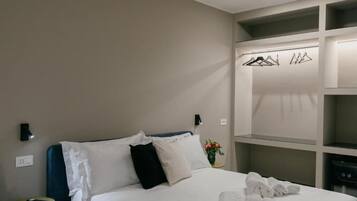 A hotel room with a bed, a bedside table with a phone, a wall-mounted light, a closet with hangers, and a vase with flowers.