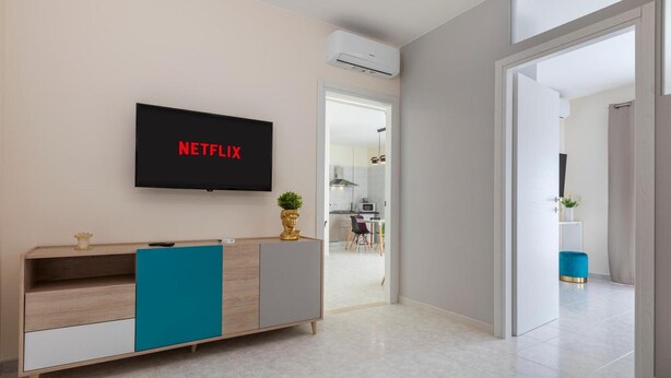 Ein modernes Wohnzimmer mit einem Fernseher, der 'NETFLIX' zeigt, einem Holzkasten und einer Tür zu einem weiteren Raum.