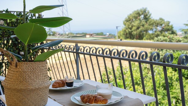 Un balcón con una mesa lista para el desayuno, una planta en maceta y vista al mar.