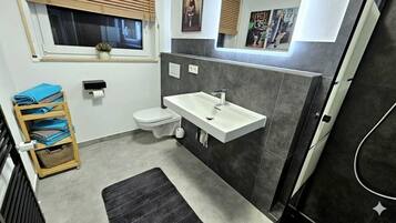 Un baño moderno con un lavamanos blanco, baldosas oscuras, un inodoro y una ventana con persianas.