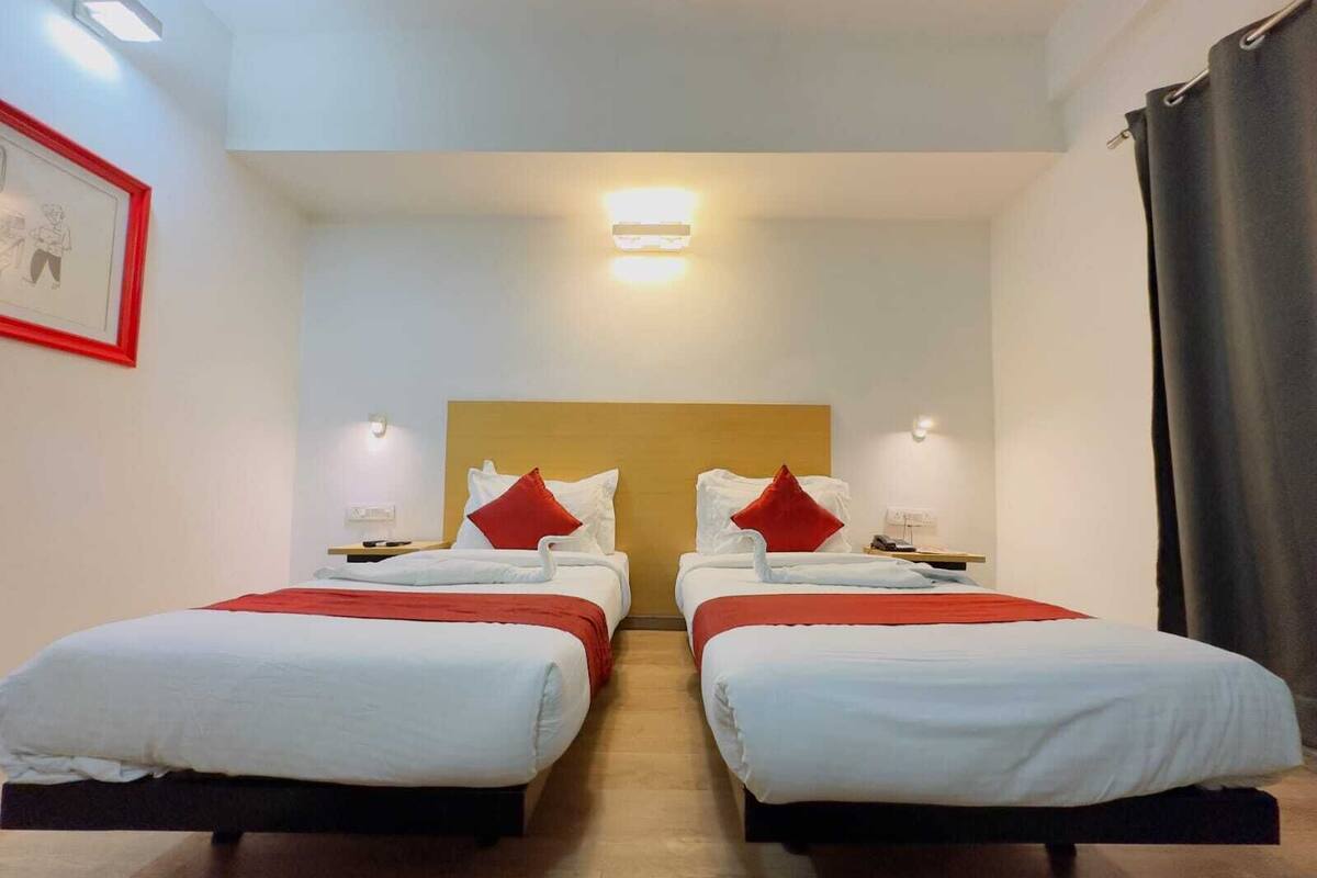 A hotel room with two beds, each with white bedding and red pillows. There are bedside tables with lamps, a wooden headboard, and a framed picture on the wall.