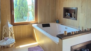 Een houten blokhut interieur met een groot bad, een klein tafeltje met een kaars en een raam met uitzicht op groen.